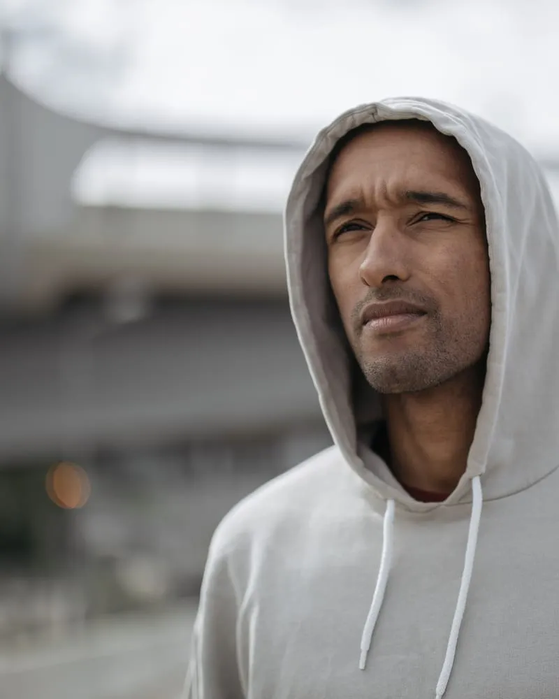 A model in a cream heavyweight pullover hoodie, hood up, against an urban concrete backdrop.