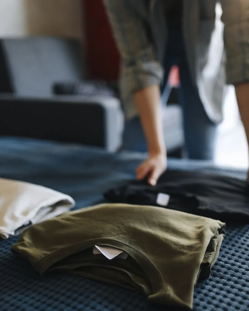 A stack of folded cotton T-shirts in olive, white and black, one showing a sewn-in woven label, ready to pack.