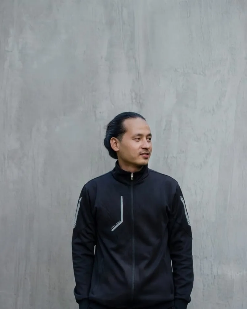 A model in a black zip track jacket with reflective sleeve stripes, against a concrete wall.